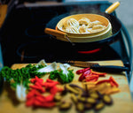 Shot of bamboo steamer on stove with steam, and dumplings inside. Colorful vegetables in foreground.  